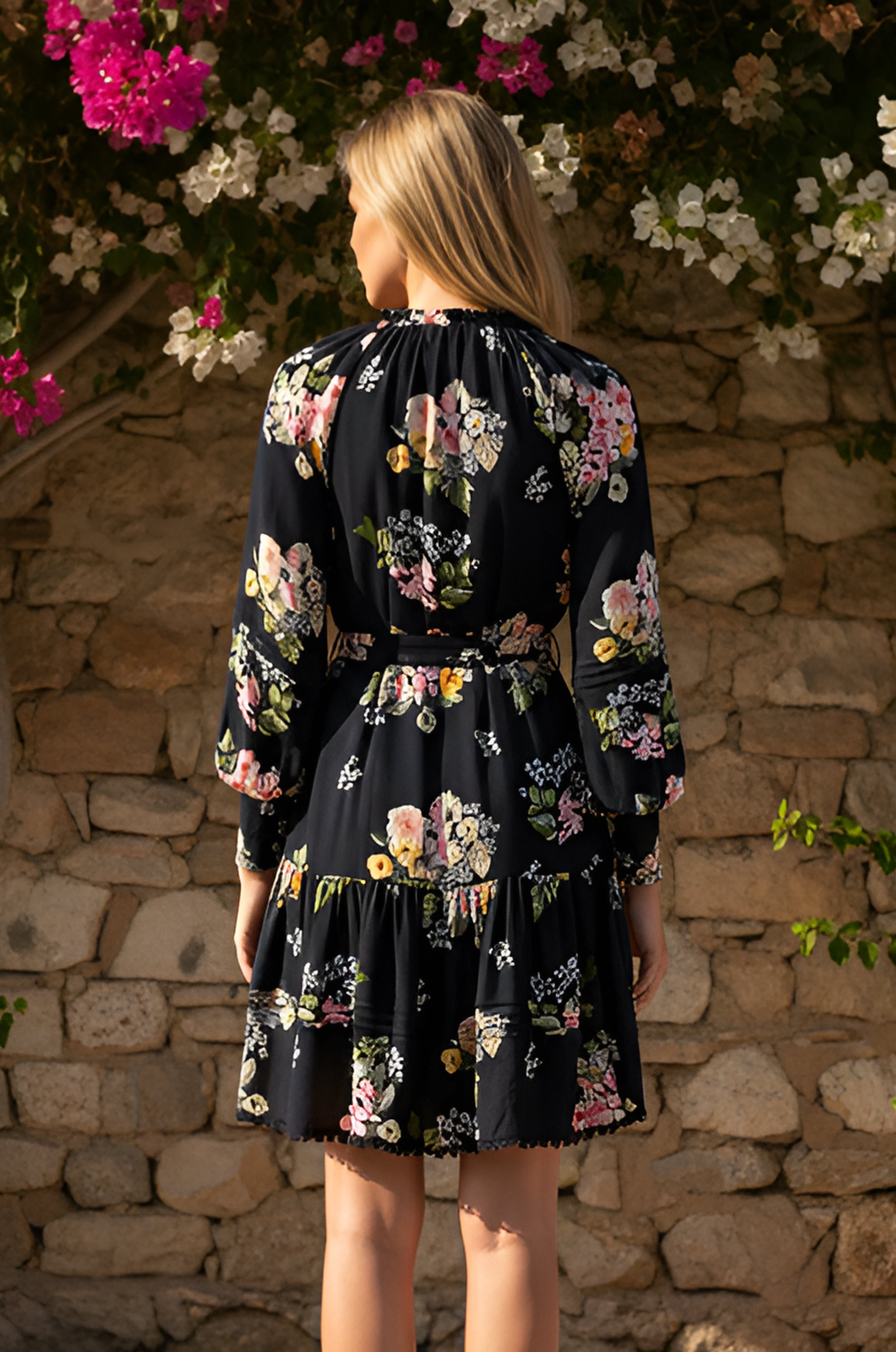A blonde woman wearing the Black Short Casual Floral Dress: Cora stands facing a stone wall with blooming flowers above.